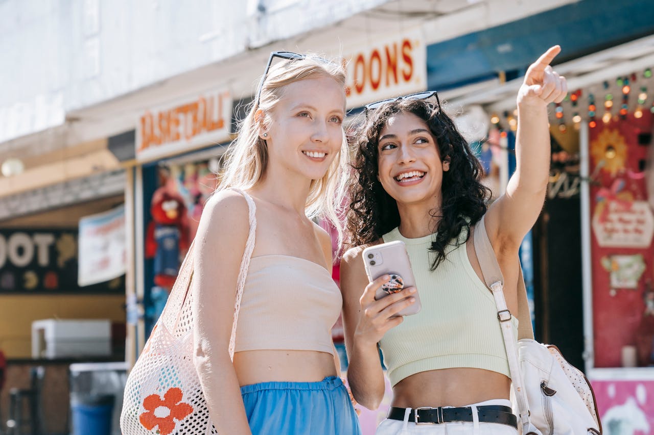 Two women smiling and pointing while shopping outdoors in a vibrant market area.